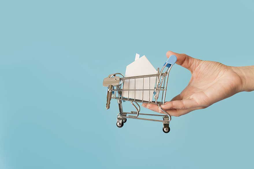 close up hand holding miniature shopping cart with paper house keys against blue background | Techlog.gr - Χρήσιμα νέα τεχνολογίας