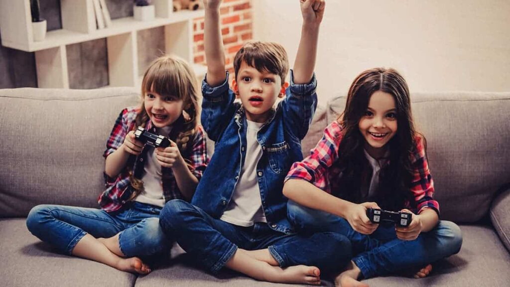 Cheerful kids are sitting together on sofa at home. Girls are playing game console while boy is cheering for them | Techlog.gr - Χρήσιμα νέα τεχνολογίας